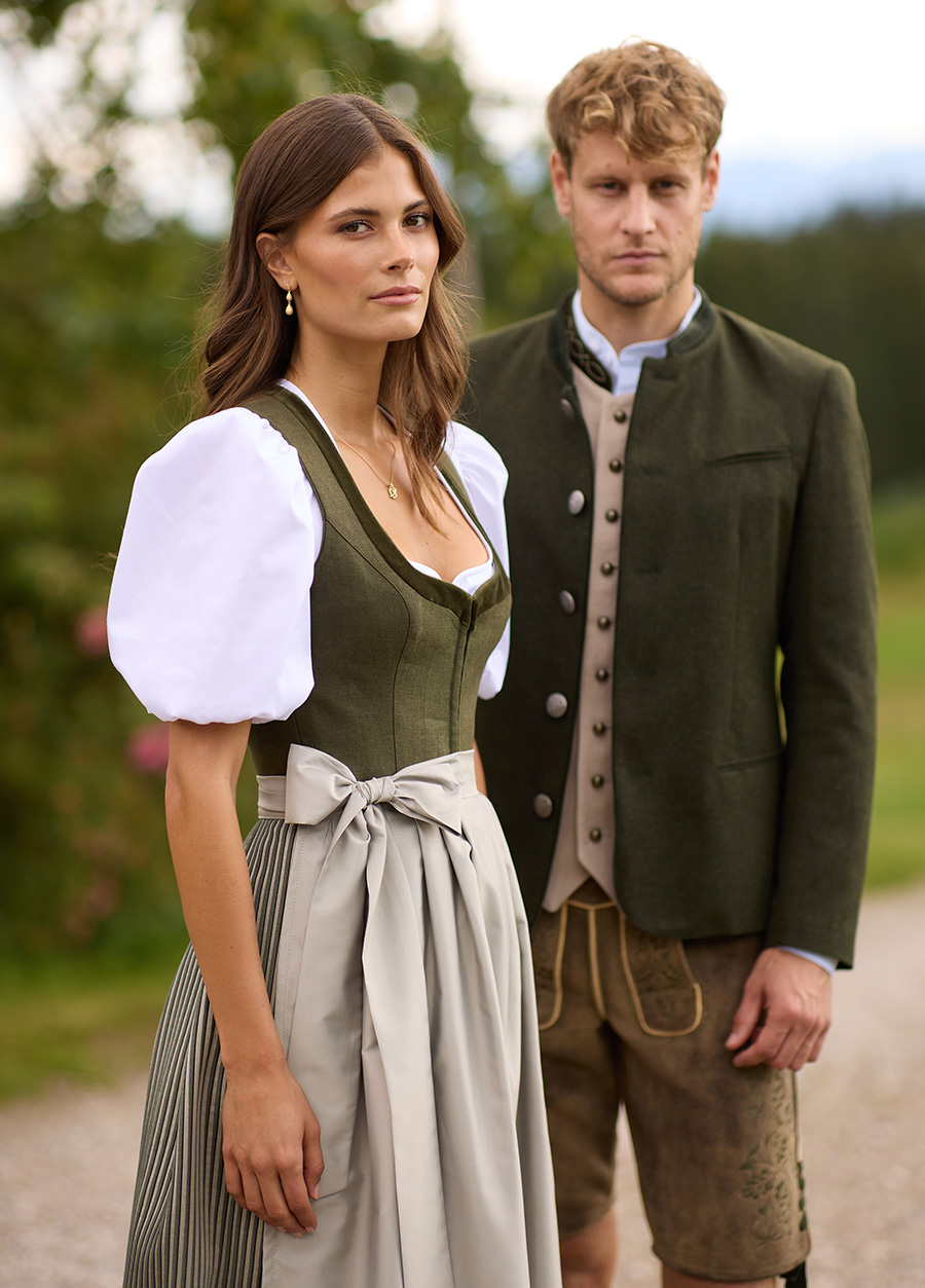 Ein Paar in elegantem Olivgrün | Das Dirndl „Isolde“ kombiniert ein olivgrünes Leinenmieder mit Samtblende, Herzausschnitt und Hakenverschluss. Ein Statement setzt der gestreifte Plisseerock. Abgerundet wird der Look durch eine schlichte, leicht glänzende Schürze sowie die Dirndlbluse „Heidi“ mit Puffärmeln. Der Janker „Josef“ in der Farbe Olive ist die perfekte Kombination aus Tradition und Stil. Hier ergänzt mit der Weste „Fridolin“, die durch ihren hochgeschlossenen, klassischen Schnitt besticht. | www.heimatglueck-tracht.de | Foto: © Heimatglück GmbH